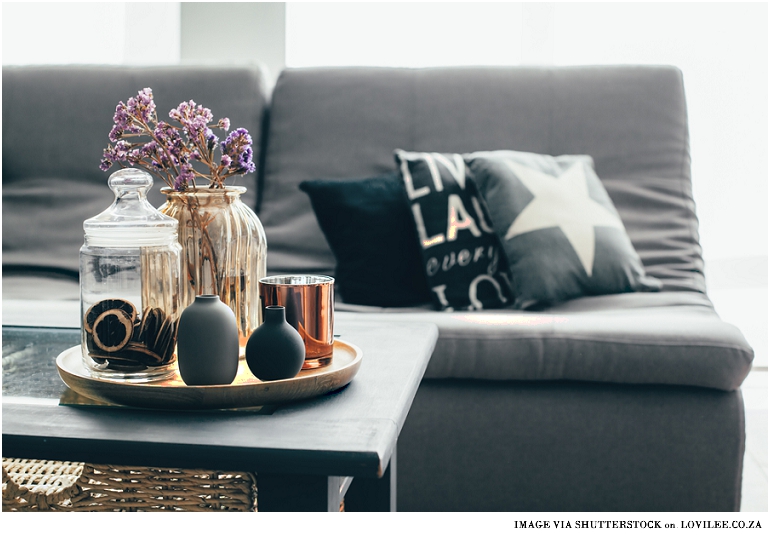 Home interior decor in gray and brown colors: glass jar with dried flowers, vase and candle on the wooden tray on the coffee table over sofa with cushions. Living room decoration. By Alena Ozerova via Shutterstock